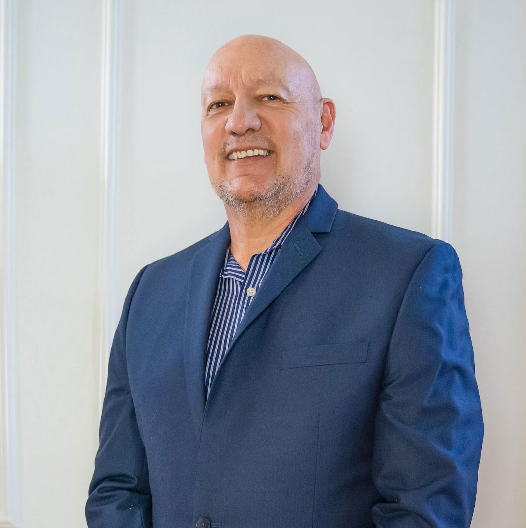 A bald man dressed in a blue suit, standing against a white wall.
